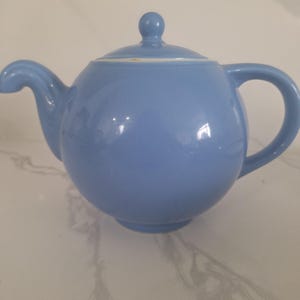 May include: A light blue ceramic teapot with a rounded body and a simple design. The teapot has a lid with a knob on top and a spout on one side.