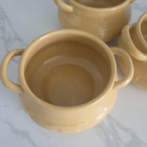 May include: Several light yellow ceramic soup bowls with handles. The bowls are round with a slightly flared rim and two curved handles on either side. The bowls are arranged on a white marble surface.