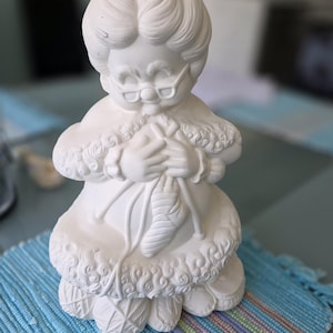 May include: A white ceramic figurine of a woman knitting. The figure wears glasses and a shawl, with yarn and knitting needles in her hands. The figurine is detailed with textured patterns and is unpainted.