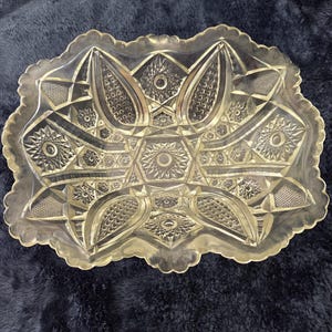 May include: Clear, decorative glass serving dish with a scalloped edge. The dish features a detailed, symmetrical pattern of geometric shapes, including stars, ovals, and diamond-like designs. The dish is likely used for serving food or as a decorative piece.