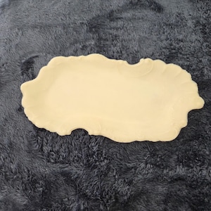 May include: A cream-colored, oblong serving tray with a scalloped edge and embossed floral detailing. The tray is made of ceramic or porcelain and is suitable for serving appetizers or desserts. The tray is approximately 12 inches long.