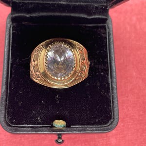 Peut inclure: Une bague dorée ornée d'une grande pierre transparente de taille ovale au centre. L'anneau est large et présente des détails gravés sur les deux côtés, incluant les chiffres 19 et 51. La bague est présentée dans un écrin doublé de velours noir.