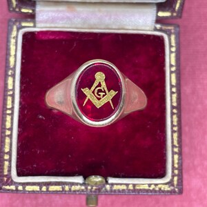 May include: Gold-toned ring with a red oval center featuring a gold Masonic symbol. The ring is displayed in a vintage, velvet-lined box with a gold clasp. The box has a gold trim and is set against a pink background.