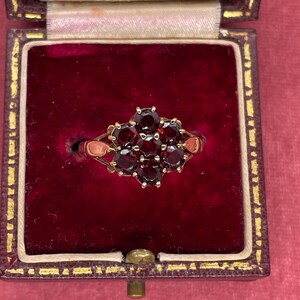 Puede incluir: Un anillo de oro con un grupo de piedras preciosas rojas oscuras de talla ovalada. El anillo se exhibe en un joyero vintage forrado de terciopelo granate con ribetes dorados. Las piedras preciosas están dispuestas en un patrón floral.