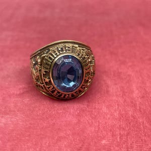 May include: A gold ring with an oval, light blue gemstone. The ring's band has detailed engravings and the words "VETERAN SCHOOL" are inscribed around the gemstone. The ring is set against a pink background.