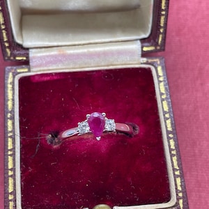 Könnte beinhalten: Ein silberner Ring mit einem birnenförmigen Rubin-Mittelstein, flankiert von zwei kleineren Diamanten. Der Ring ist in einem klassischen Design gehalten und wird in einer Vintage-Schmuckschatulle mit rotem Samt präsentiert.