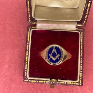 Könnte beinhalten: Ein goldener Ring mit blau emaillierter Oberfläche, der ein Freimaurersymbol zeigt, präsentiert in einer mit weinrotem Samt ausgekleideten Schmuckschatulle. Der Ring hat eine abgerundete Oberfläche und ein poliertes Goldband.