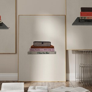 May include: A framed print showing a stack of books and a decorative box. The books include titles such as "Film for Her" and "Funny Weather." The box is rectangular with a patterned design. The frame has a gold border.