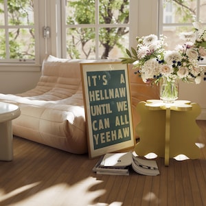 Peut inclure: Un poster encadré avec le texte "IT'S A HELLNAW UNTIL WE CAN ALL YEEHAW" est posé à côté d'une table d'appoint jaune et d'un bouquet de fleurs dans un vase en verre. Un canapé beige et une pile de livres sont également visibles.