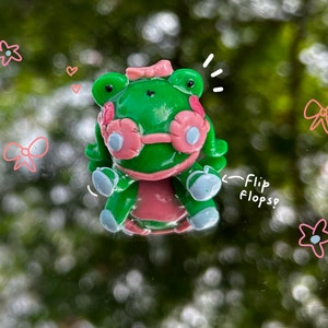 Puede incluir: Una figurita de rana verde con detalles rosas, que lleva un lazo rosa y gafas de sol. La rana está sentada sobre una superficie transparente con un fondo verde borroso. El texto "Flip Flops?" está escrito en negro sobre la superficie.
