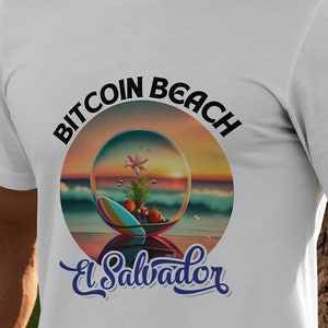Könnte beinhalten: Weißes T-Shirt mit einem schwarzen und weißen Kreis-Motiv, auf dem "Bitcoin Beach El Salvador" steht. Das Motiv zeigt eine Strandszene in einer Glaskugel.