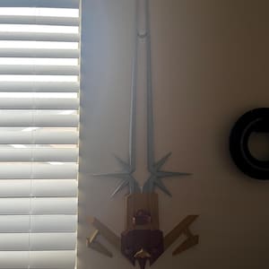 Op de afbeelding: Een decoratief zilveren en gouden zwaard met een rode handgreep gemonteerd op een witte muur. Het zwaard heeft een stervormig ontwerp aan de boven- en onderkant.