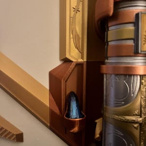 Op de afbeelding: Close-up van een 3D-geprint wandbevestiging met een decoratieve gouden en koperen afwerking. De bevestiging heeft een klein, blauw kristal in een ingezonken nis. De bevestiging is ontworpen om een decoratief object te houden, zoals een zwaard of een staf.