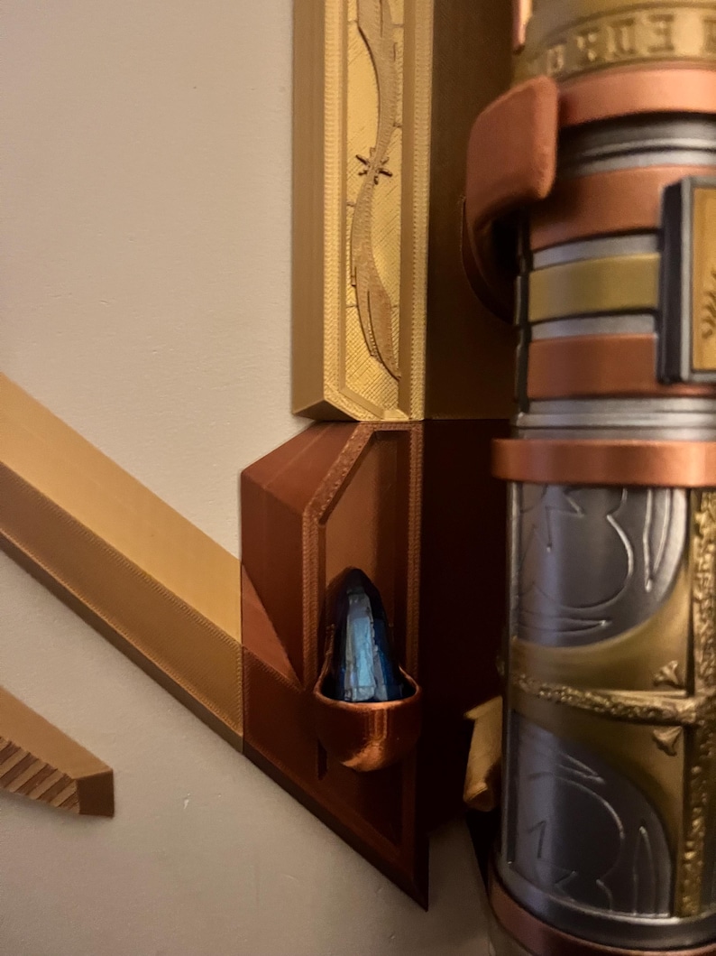 Op de afbeelding: Close-up van een 3D-geprint wandbevestiging met een decoratieve gouden en koperen afwerking. De bevestiging heeft een klein, blauw kristal in een ingezonken nis. De bevestiging is ontworpen om een decoratief object te houden, zoals een zwaard of een staf.
