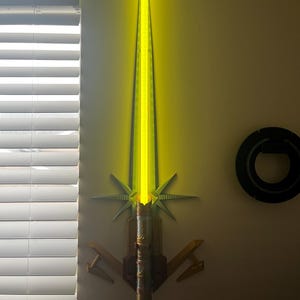 Op de afbeelding: Een gele lightsaber met een gouden handvat en een decoratieve standaard. De lightsaber is aan de muur gemonteerd.