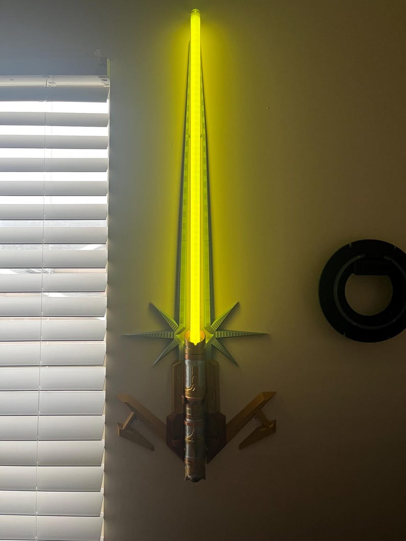 Op de afbeelding: Een gele lightsaber met een gouden handvat en een decoratieve standaard. De lightsaber is aan de muur gemonteerd.