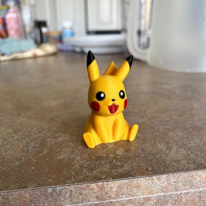 May include: A yellow Pikachu figurine with black-tipped ears, red cheeks, and a red tongue. The figurine is sitting on a brown countertop. The Pikachu figure is a popular character from the Pokémon franchise.