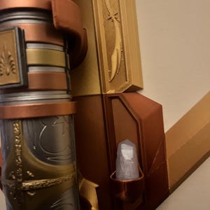 Op de afbeelding: Een 3D-geprinte wandbevestiging met een gouden en koperen afwerking. De bevestiging heeft een decoratief ontwerp en een klein vakje voor een kristal. Het kristal is helder en heeft een licht troebele uitstraling.