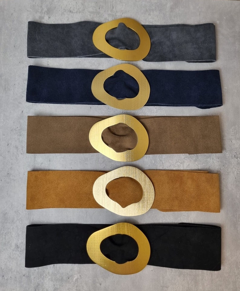 May include: Five suede belts with large gold oval buckles. The belts are in different colors: gray, navy blue, brown, tan, and black.