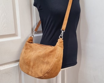 May include: A tan suede shoulder bag with a matching adjustable strap. The bag is displayed on a mannequin wearing a black top. The bag has a curved shape and silver-tone hardware.