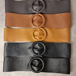 May include: Four leather belts with round, oversized buckles. The belts are in different colors: brown, tan, black, and dark gray.