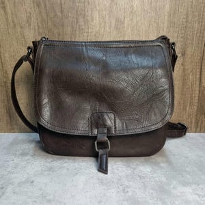May include: A brown leather crossbody bag with a flap closure and adjustable strap. The bag has a textured surface and a buckle detail on the front. The bag is designed with a zip closure at the top.