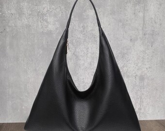 May include: A black hobo bag with a pebbled texture, featuring a wide, triangular silhouette, a single integrated shoulder strap, and a small silver-toned metal zip pull visible on the upper side of the bag's opening.