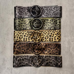 Puede incluir: Cinco cinturones de estampado de leopardo con hebillas redondas de metal. Los cinturones son de diferentes tonos de marrón, negro y verde.