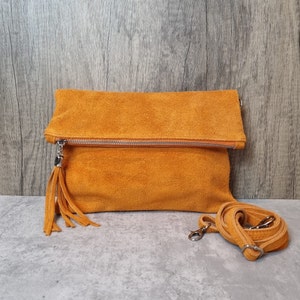 May include: A mustard yellow suede clutch bag with a long strap and a tassel. The bag has a zipper closure and a flap.
