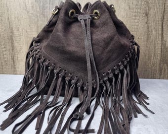 May include: A brown suede bucket bag with long fringe detailing. The bag has a drawstring closure and gold-toned metal eyelets. The fringe hangs from the bottom edge of the bag, adding a bohemian flair. The bag is made of a soft, textured material.