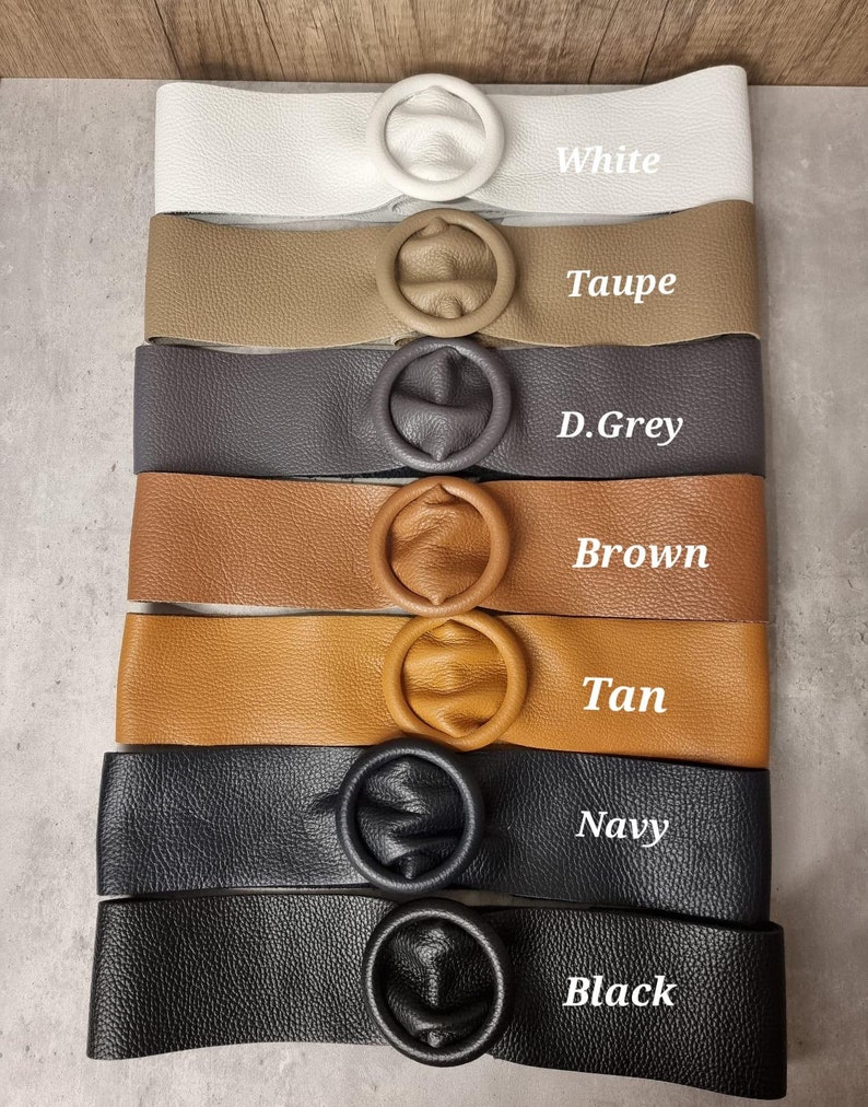May include: A stack of eight leather belts with round metal buckles in various colors, including white, taupe, dark grey, brown, tan, navy, and black. The belts are arranged vertically with the buckle facing the viewer. Each belt has a label with the color name written below it.