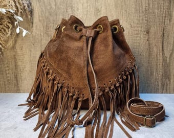 May include: A brown suede bucket bag with long fringe detailing. The bag has a drawstring closure and a matching adjustable shoulder strap. The bag is a warm brown color and has a bohemian style.