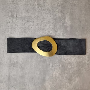 May include: A black suede belt with a large gold oval buckle. The buckle has a cutout in the center.