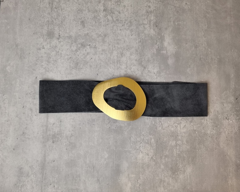 May include: A black suede belt with a large gold oval buckle. The buckle has a cutout in the center.