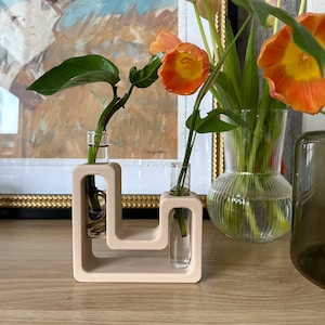 Puede incluir: Un jarrón de madera beige claro con dos tubos de ensayo de vidrio para sostener flores. El jarrón tiene un diseño único con dos aberturas para los tubos de ensayo. El jarrón está sobre una superficie de madera con una pintura y un jarrón de flores en el fondo.