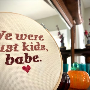 May include: A white embroidery hoop with the text "We were just kids, babe." in brown thread and a small red heart.