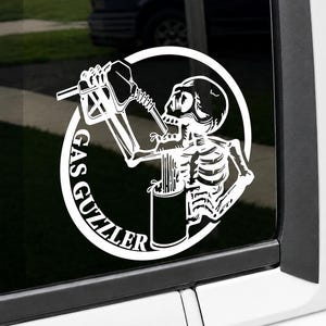 May include: A white vinyl decal featuring a skeleton holding a gas pump nozzle to its mouth and a mug in its other hand. The design is enclosed in a circular border with the text GAS GUZZLER printed along the left side in a bold font.