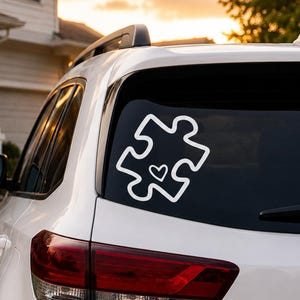 May include: A white vinyl decal applied to the rear window of a white SUV. The decal features a bold white outline of a single puzzle piece with a small, simple heart shape centered within the puzzle piece design, creating a clean graphic.