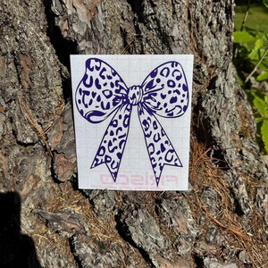 Peut inclure: Un autocollant en vinyle violet fonc&eacute; repr&eacute;sentant un n&oelig;ud avec un motif l&eacute;opard. L'autocollant est appliqu&eacute; sur une feuille de support rectangulaire blanche avec un quadrillage l&eacute;ger et un petit logo rouge imprim&eacute; au centre en bas.