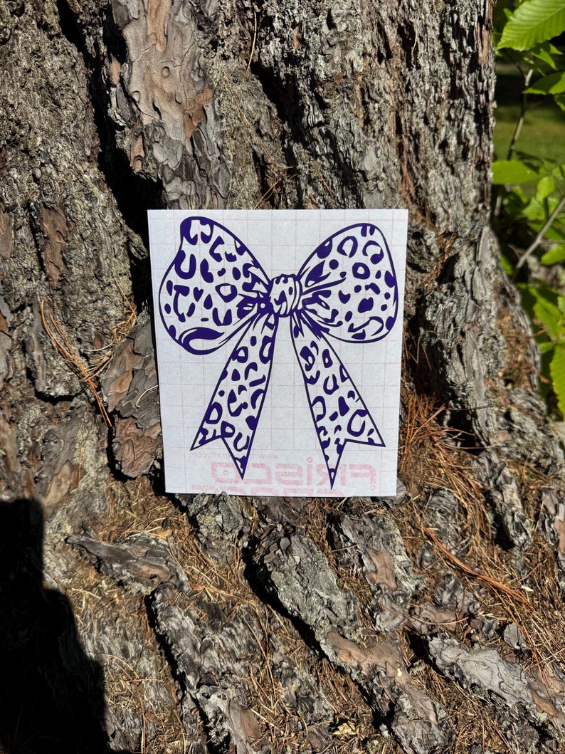 Peut inclure: Un autocollant en vinyle violet fonc&eacute; repr&eacute;sentant un n&oelig;ud avec un motif l&eacute;opard. L'autocollant est appliqu&eacute; sur une feuille de support rectangulaire blanche avec un quadrillage l&eacute;ger et un petit logo rouge imprim&eacute; au centre en bas.