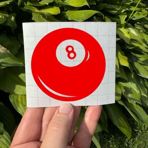 May include: A red vinyl decal featuring a circular design with a white center circle containing the number 8. The decal is applied to a white square backing sheet with a light red grid pattern, designed for application as a sticker or graphic.