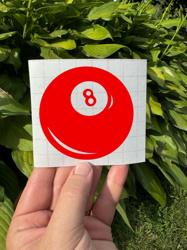 May include: A red vinyl decal featuring a circular design with a white center circle containing the number 8. The decal is applied to a white square backing sheet with a light red grid pattern, designed for application as a sticker or graphic.
