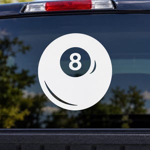 May include: A white vinyl decal of an eight ball from a billiards game, featuring a circular design with a central dark circle containing the number eight, applied to the rear window of a dark-colored vehicle with a visible red brake light.
