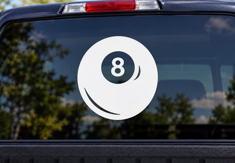 May include: A white vinyl decal of an eight ball from a billiards game, featuring a circular design with a central dark circle containing the number eight, applied to the rear window of a dark-colored vehicle with a visible red brake light.