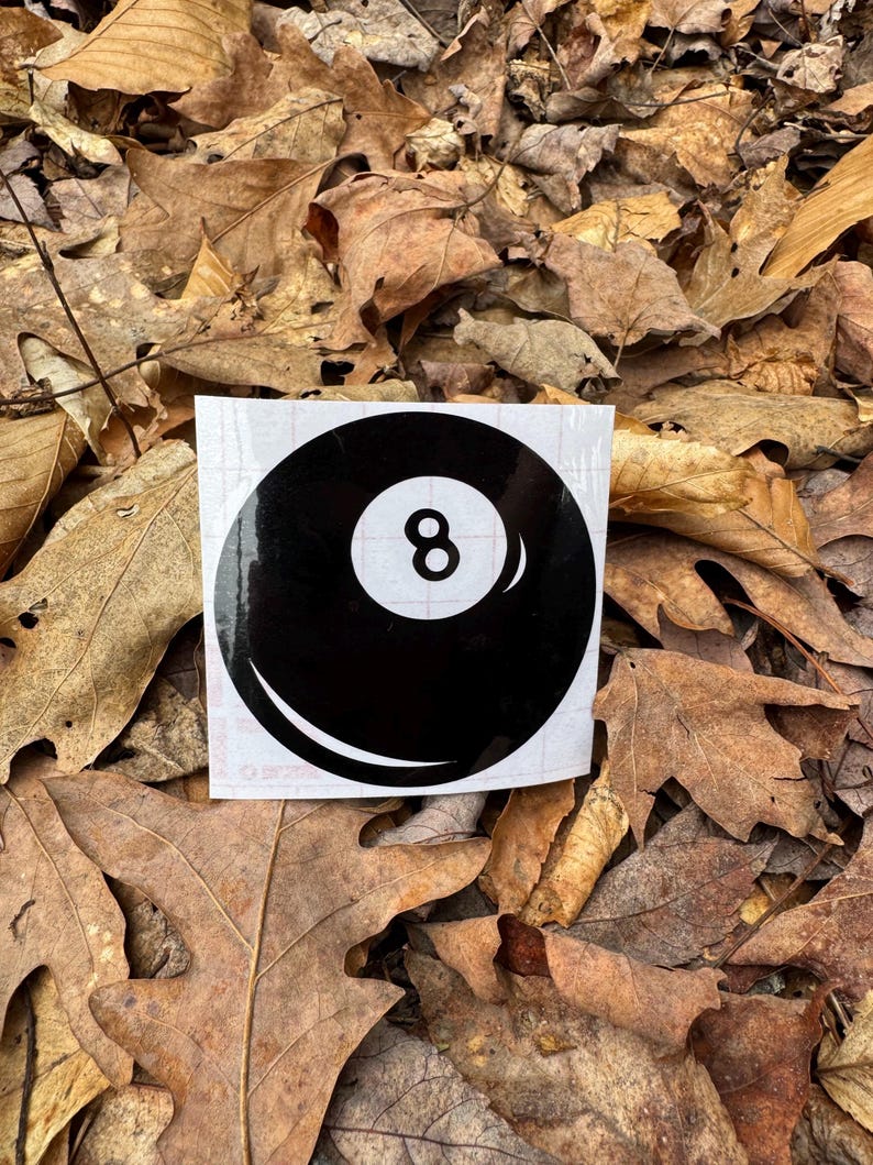 May include: A black and white vinyl decal sticker featuring the iconic design of a round billiard eight ball. The graphic displays a solid black circular shape with a central white circle containing the black number eight and a white highlight.