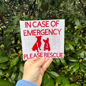May include: A white sticker with red text that reads "IN CASE OF EMERGENCY PLEASE RESCUE." The sticker features two red dog silhouettes and the numbers 1 and 2. The sticker is held in front of a background of green leaves.