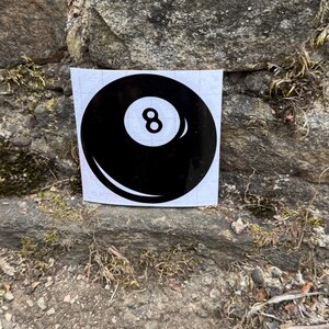 May include: A square white sticker featuring a black billiard ball graphic with a central white circle containing the number 8. The design includes a curved white highlight on the lower left and a smaller highlight on the right side of the ball.