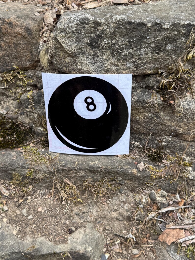 May include: A square white sticker featuring a black billiard ball graphic with a central white circle containing the number 8. The design includes a curved white highlight on the lower left and a smaller highlight on the right side of the ball.
