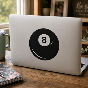 May include: A silver laptop featuring a black and white circular sticker of an eight ball with the number eight centered in a white circle. The sticker is applied to the center of the laptop lid, displaying a classic billiard ball graphic design.