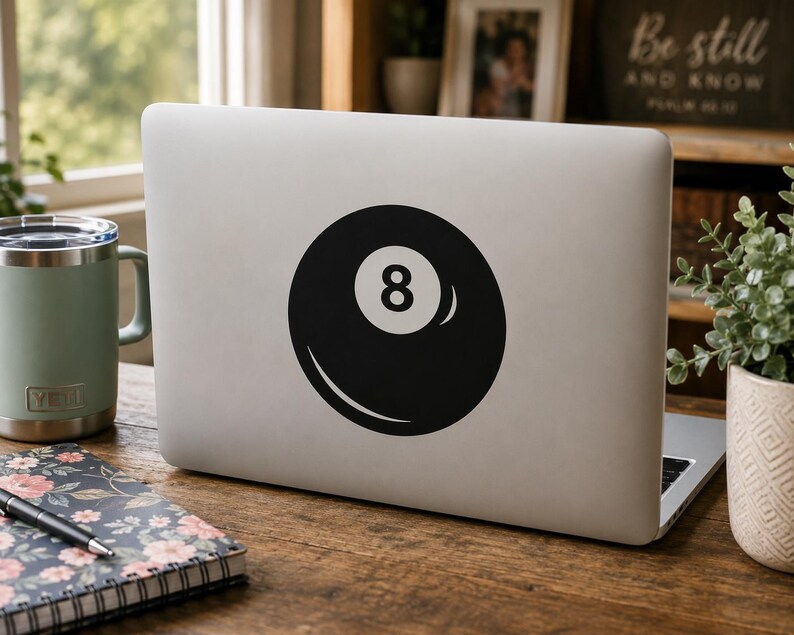 May include: A silver laptop featuring a black and white circular sticker of an eight ball with the number eight centered in a white circle. The sticker is applied to the center of the laptop lid, displaying a classic billiard ball graphic design.